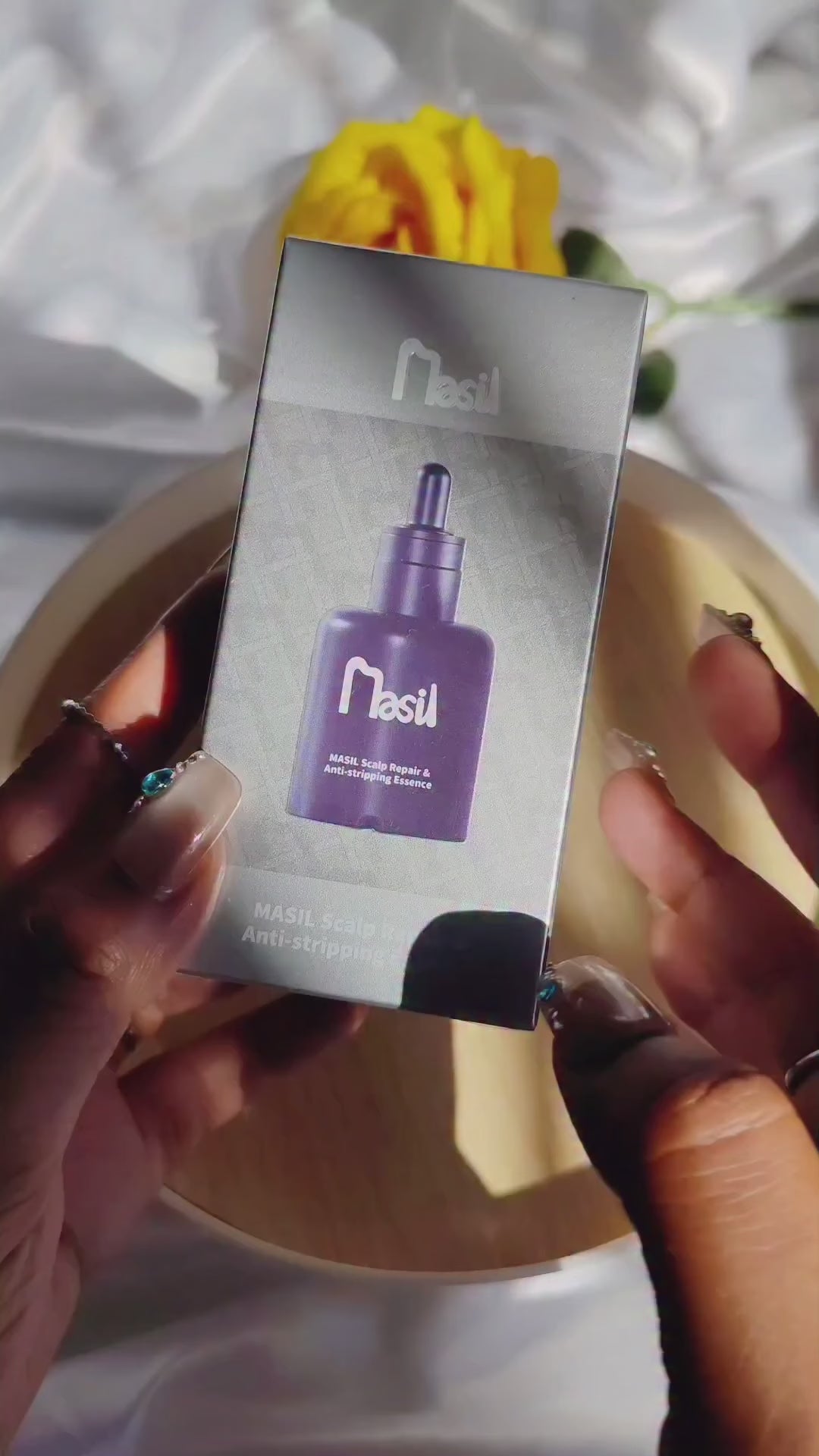 Close-up video of MASIL Scalp Repair & Anti-stripping Essence showing manicured hands unwrapping the silver packaging, revealing a matte black dropper bottle, and displaying a clear essence texture with the bottle and box styled together on a wooden tray.
